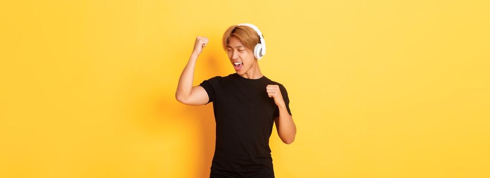 Carefree attractive asian guy with blond hair, singing along and dancing as listening to music in wireless headphones, standing yellow background.