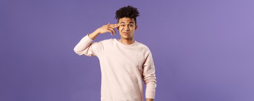 Portrait of young teenage guy, hispanic male student losing his mind at home lockdown, pointing at temple with finger gun and grimacing as shooting himself from boredom or annoyance.