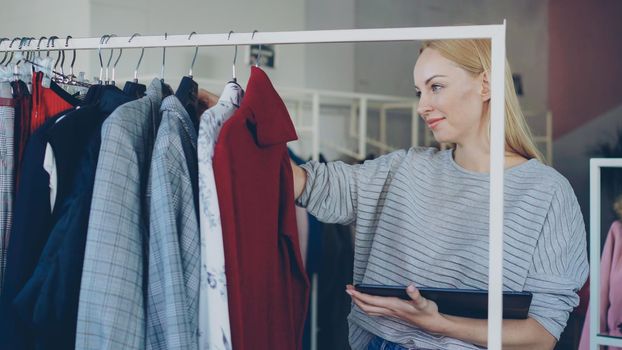 Young businesswoman is checking garments on rails and working with tablet in her clothing shop. She is checking prices and labels, touching screen and typing. Successful start-up concept.