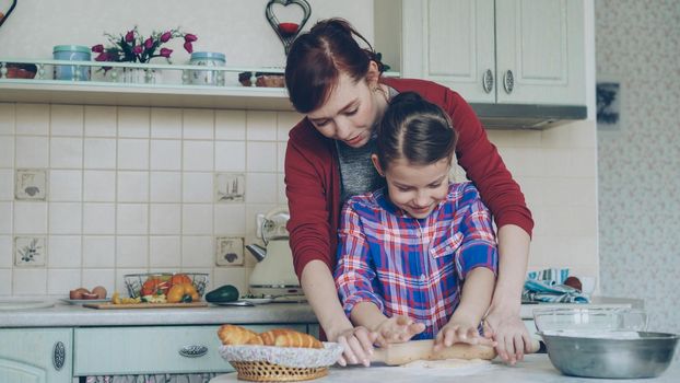 Young attractive mother teaching her little cute daughter rolling dough while cooking together in the kitchen at home on holidays. Family, food and people concept