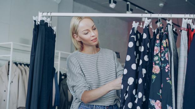 Pretty blond girl is choosing fashionable clothes in boutique. She is looking through fine garments and taking polka-dotted dress from the rails, examining its quality and length.