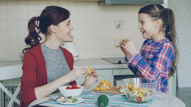 Happy family of young mother and cute cheerful daughter have fun grimacing silly with vegetables while cooking in the kitchen at home