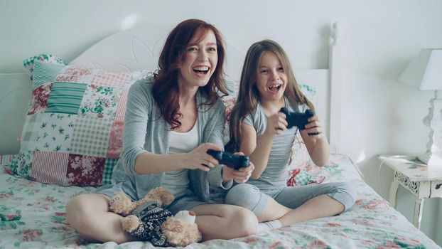 Funny girl with happy young mother have fun while playing computer console games on TV sitting on bed at home in the morning in cozy bedroom. Daughter embracing mom after winning