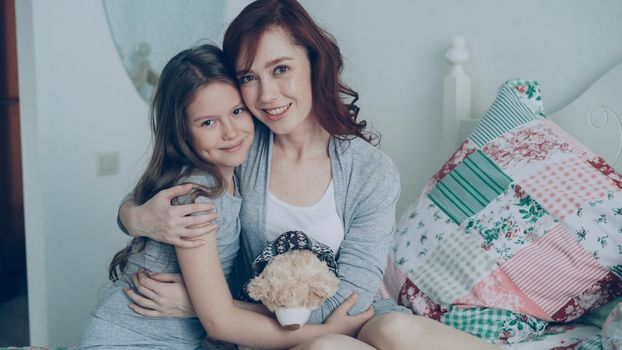 Portrait of adorable smiling girl embracing her happy mother and looking at camera together while sitting on bed in bright cozy bedroom at home