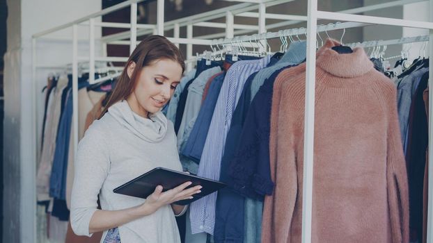 Young and successful owner of women's clothing shop is checking and counting garments on rails while using tablet. She is typing information about her goods. Small business concept.