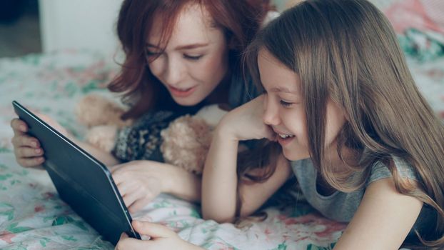 Close-up of attractive young mother and her smiling daughter in pajamas laughing and looking in digital tablet while lying on bed at home