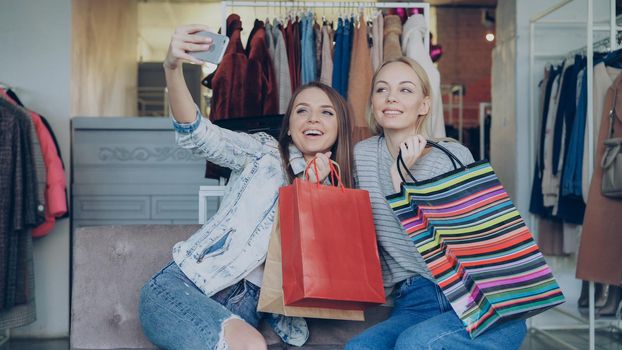 Cute girls are sitting and making selfie with colourful paper bags using smartphone in luxurious women's clothing boutique, then watching pictures together. They are smiling and laughing carelessly.