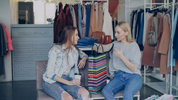 Two young women discussing purchased ankle shoes while having rest in expensive boutique. Girls are excited about shopping, gestiuring and laughing