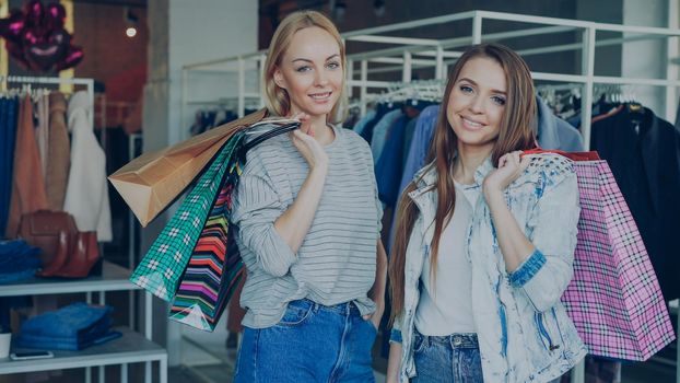 Portrait of beautiful young women are standing with lots of paper bags in their hands in luxurious women's clothes boutique. Fashionable clothes, bags, shoes and baloons in background.