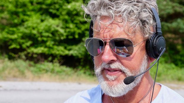 Gray-haired elderly man freelancer with a beard in sunglasses, working in the headphones on nature. A crazy and extraordinary old senior in the summer