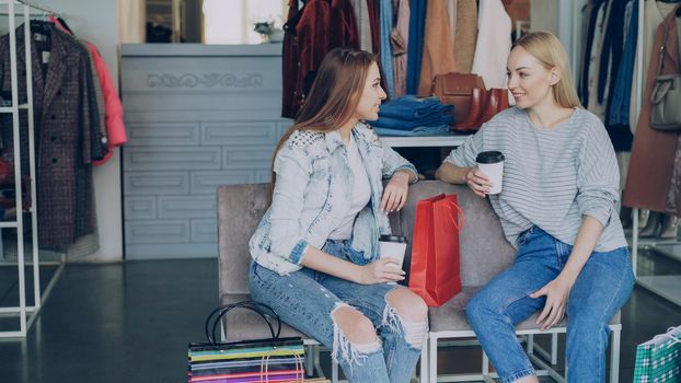 Two young attractive ladies sitting in clothes department in mall and talking happily after shopping. Colourful women's garments and bags in background.