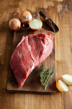 A big chunk of red met on vintage cutting board next to white onions. Green rosemary. Pepper beans.