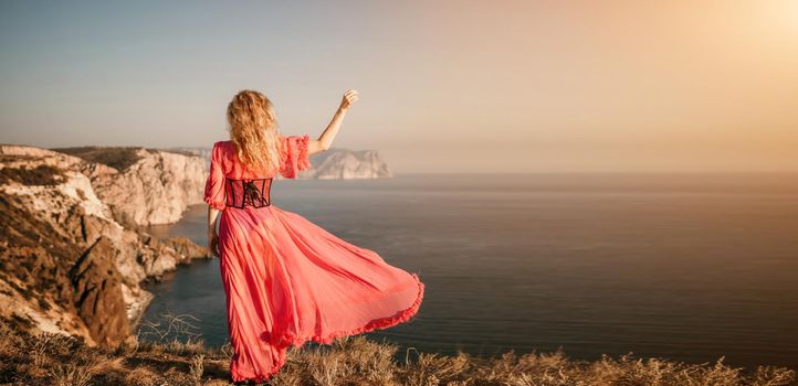 Side view a Young beautiful sensual woman in a red long dress posing on a volcanic rock high above the sea during sunset. Girl on the nature on blue sky background. Fashion photo