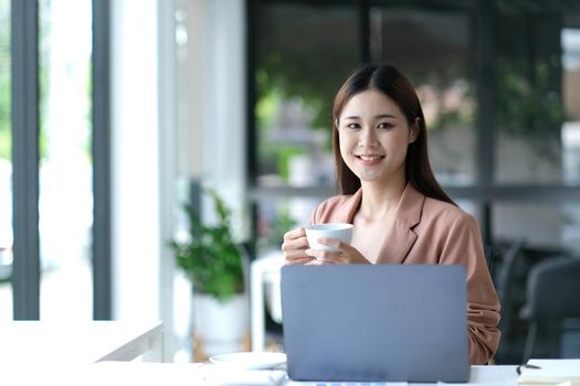 Smiling Asian businesswoman holding a coffee mug and laptop at the office. Looking at the camera..