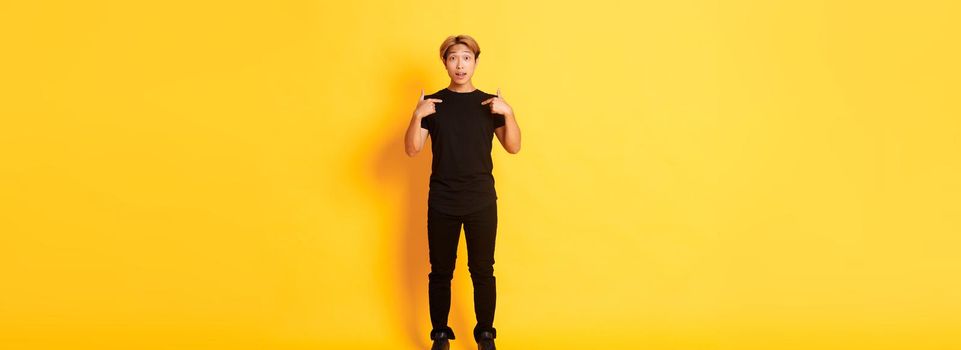 Full-length of surprised attractive korean blond guy in black outfit pointing at himself impressed, yellow background.