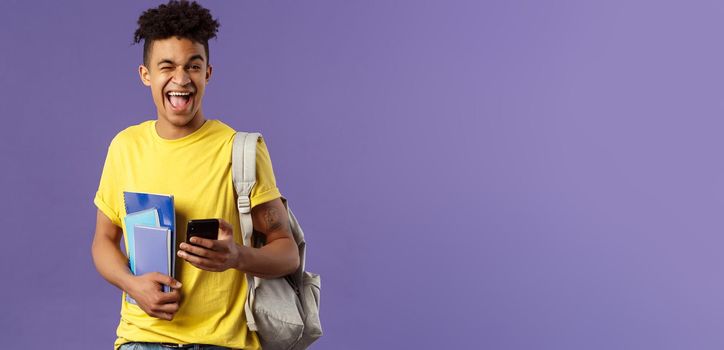 Back to school, university concept. Portrait of joyful, smiling happy man, student using mobile phone, wink at camera upbeat, holding backpack and notebooks, notes from lecture and smartphone.