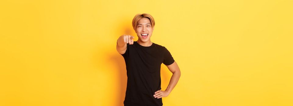 Portrait of cheerful handsome asian guy with blond hair choosing you, smiling and pointing finger at camera, congrats gesture.