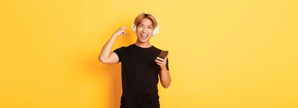 Happy satisfied asian guy likes music or podcast, pointing finger at headphones, holding smartphone, standing yellow background.