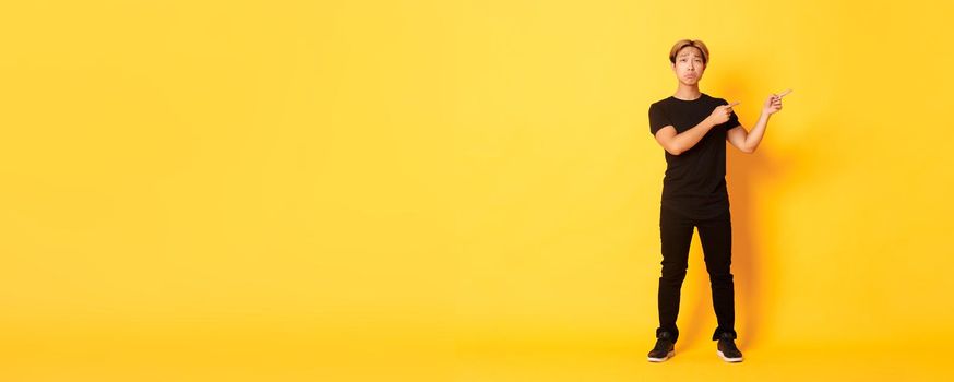 Full-length of sad and miserable asian guy in black clothes, pouting, pointing fingers upper right corner, standing yellow background.