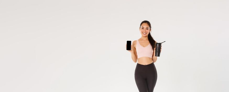 Full length of smiling good-looking asian fitness girl, sportswoman in activewear showing smartphone screen and water bottle, control drinking liquid with mobile application during training session.