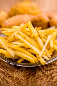 Raw cut french fries in a transparent glass plate along with raw potato with it on jute bag's surface.