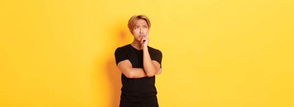 Indecisive moody asian guy sulking, looking away thoughtful, standing over yellow background, making decision.