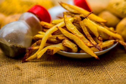 Fresh fired French fries of along with sprinkler of black pepper and salt on jute bag's surface and some raw potato also.