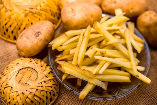 Raw cut french fries in a transparent glass plate along with raw potato with it on jute bag's surface.