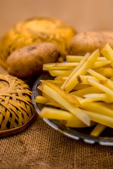 Raw cut french fries in a transparent glass plate along with raw potato with it on jute bag's surface.