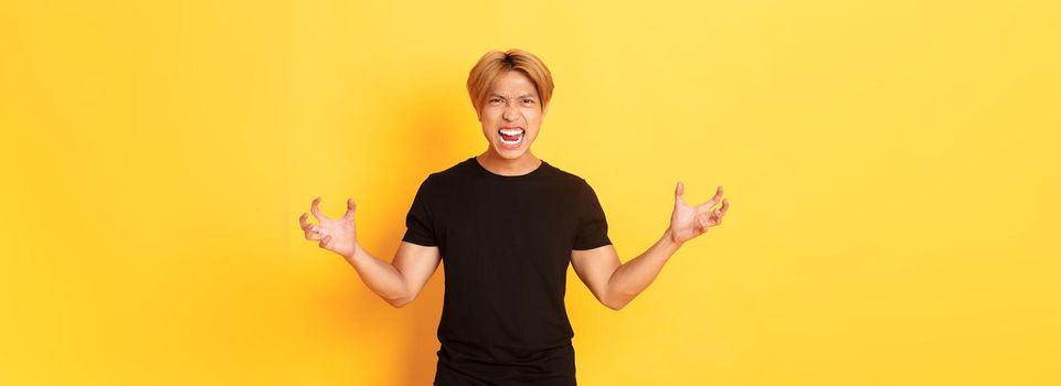Portrait of outraged, pissed-off asian man shaking hands hateful and grimacing, shouting angry, standing over yellow background.