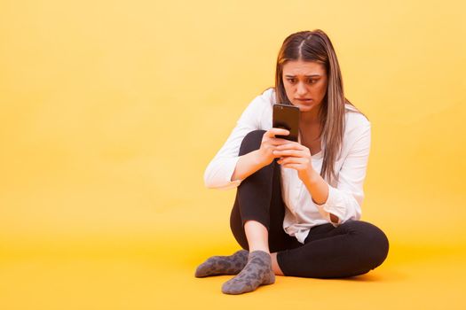 Girl looking worried at her phone while sitting down over yellow background. Facial expression