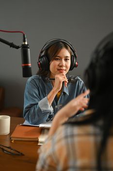 Positive female radio host listening to interesting conversation with guest during recording podcast in broadcasting home studio.