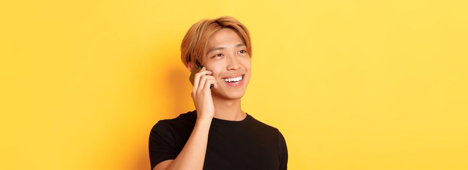 Close-up of handsome carefree asian guy with fair hair, talking on mobile phone and smiling happy, standing yellow background.