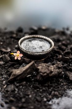 Close up of activated charcoal in a glass bowl on the wooden surface along with some raw powder of charcoal or coal spread on the surface.