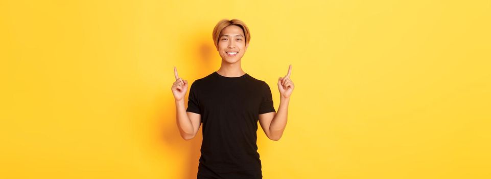 Portrait of handsome smiling asian guy in black t-shirt pointing fingers up and looking satisfied, yellow background.