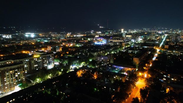 View from the height of the night city of Almaty. Different lights are shining brightly. Cars are driving on the roads. The Kok-Tobe TV tower is visible in the distance. Lights are burning in windows