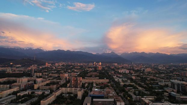 Epic sunset with clouds over the city of Almaty. The gradient of clouds is from dark blue to purple-orange. Green tall trees, houses, road with cars. The lights are on. High mountains in the distance
