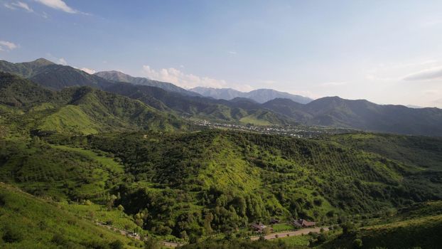 The mountains are covered with green trees and grasses. Top view from the drone on the road with cars, there are houses in places. A small town is visible in the distance. A green forest is growing