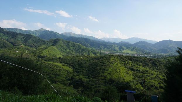 High green mountains and a small town in the distance. Christmas trees and green grass grow. There are white clouds in the sky. The shadow of the clouds falls on the hills. A wire has been conducted