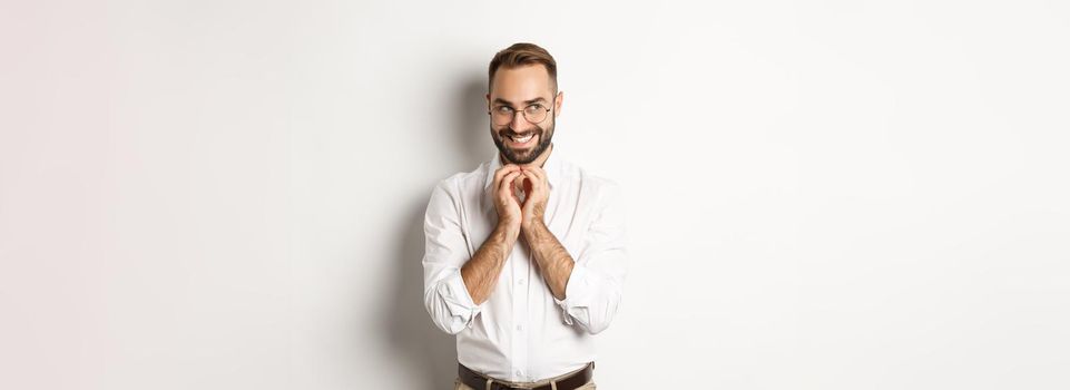 Young cunning businessman steeple fingers and smiling devious, scheming plan, standing over white background.