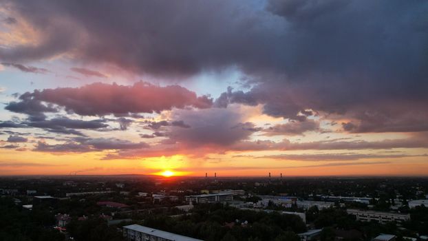 Epic sunset with clouds over the city of Almaty. The gradient of clouds is from dark blue to purple-orange. Green tall trees, houses, a road with cars. The lights are on. The sun goes below horizon