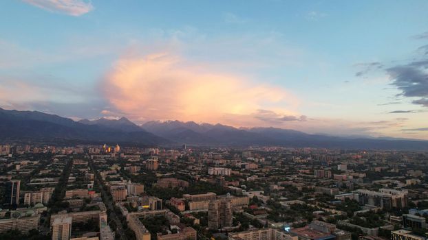 Epic sunset with clouds over the city of Almaty. The gradient of clouds is from dark blue to purple-orange. Green tall trees, houses, road with cars. The lights are on. High mountains in the distance