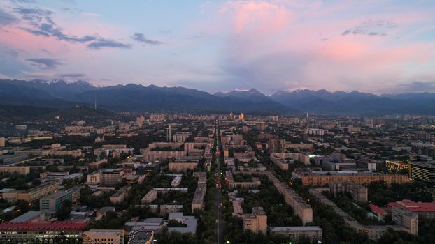 Epic sunset with clouds over the city of Almaty. The gradient of clouds is from dark blue to purple-orange. Green tall trees, houses, road with cars. The lights are on. High mountains in the distance