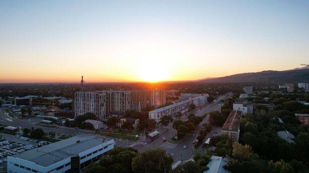 Dawn over the tall houses and the city of Almaty. Cars are driving on the road. There are many green trees growing. Houses are being built. Green hills are visible in distance. Sun rays fall on city