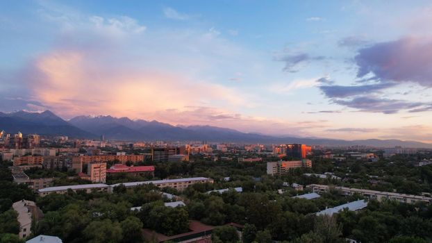 Epic sunset with clouds over the city of Almaty. The gradient of clouds is from dark blue to purple-orange. Green tall trees, houses, road with cars. The lights are on. High mountains in the distance