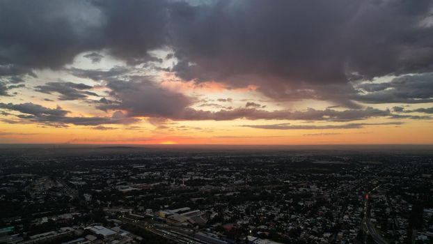 Large gray clouds in the orange light of the sun at sunset over the city of Almaty. A large city with low houses. Cars are driving on the road, lights are on. Green trees are growing. Drone view