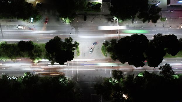 View from the height of the night intersection. Bright lights from cars, a lot of traffic on the road. Several cars are waiting for their turn to pass. Lanterns are lit. There are green trees. Almaty