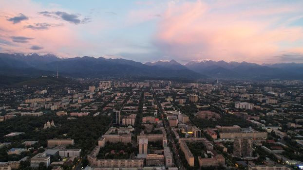 Epic sunset with clouds over the city of Almaty. The gradient of clouds is from dark blue to purple-orange. Green tall trees, houses, road with cars. The lights are on. High mountains in the distance