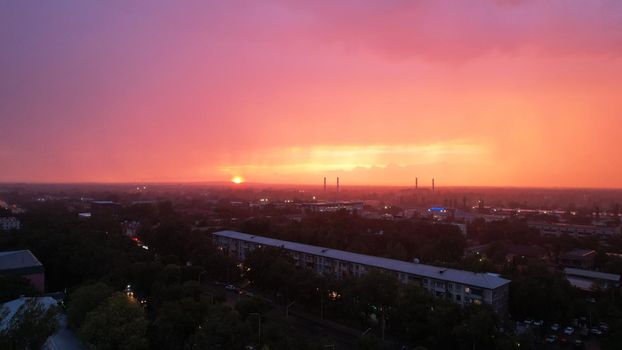 Epic sunset with clouds over the city of Almaty. The gradient of clouds is from dark blue to purple-orange. Green tall trees, houses, a road with cars. The lights are on. It's raining heavily