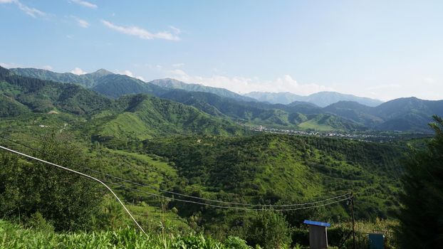 High green mountains and a small town in the distance. Christmas trees and green grass grow. There are white clouds in the sky. The shadow of the clouds falls on the hills. A wire has been conducted